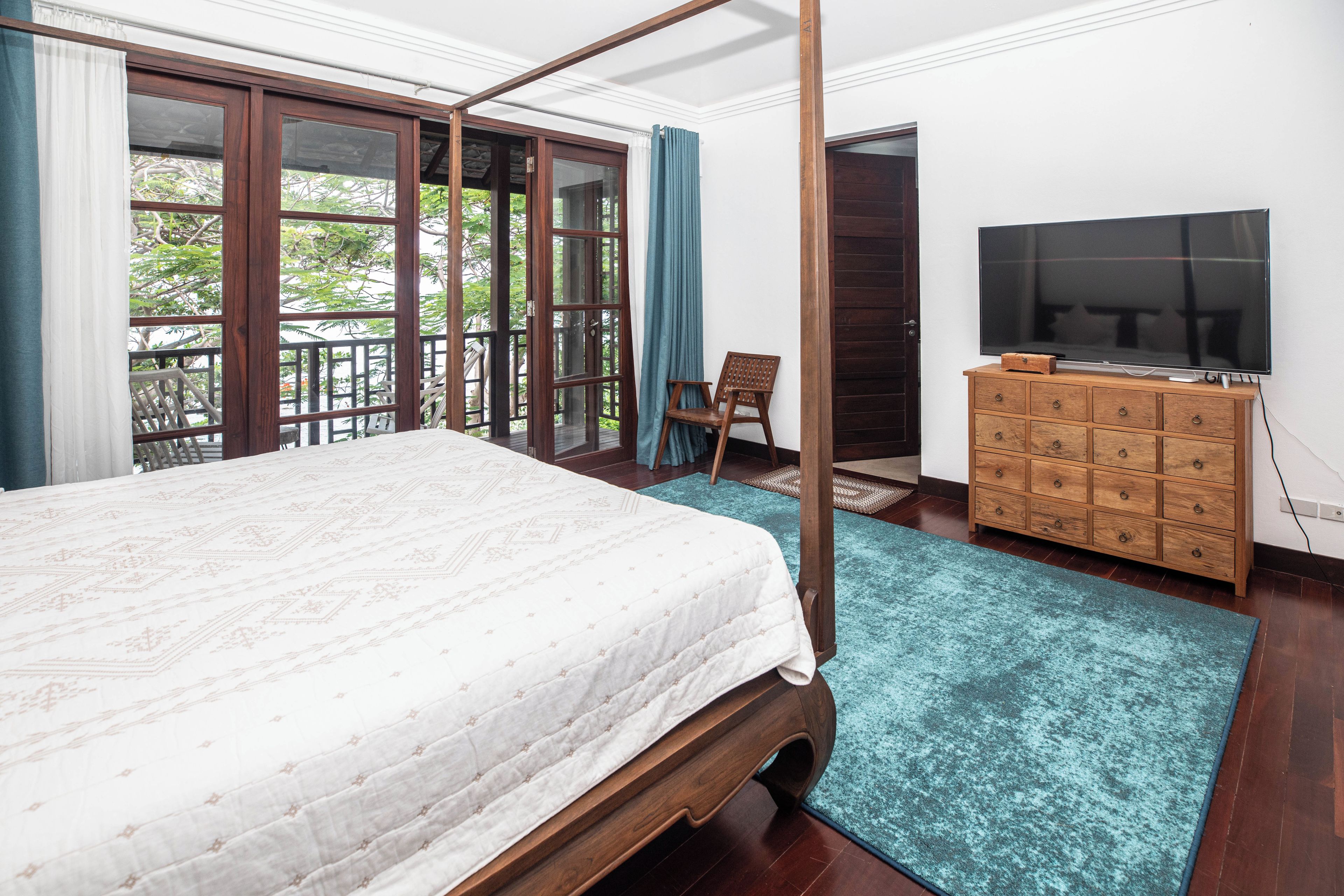 Un dormitorio con cama con dosel, un televisor sobre un aparador de madera y un balcón con vistas a árboles.