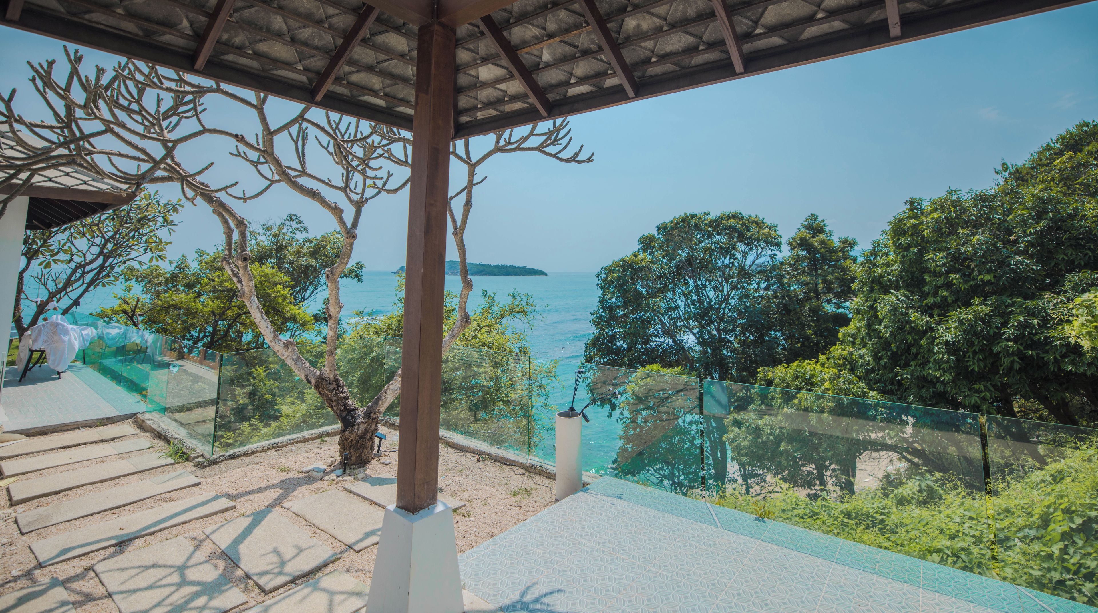 A covered outdoor area with a view of the sea, a tree, and a glass railing.