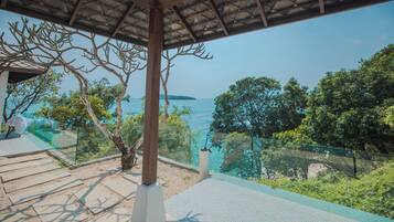 A covered outdoor area with a view of the sea, a tree, and a glass railing.