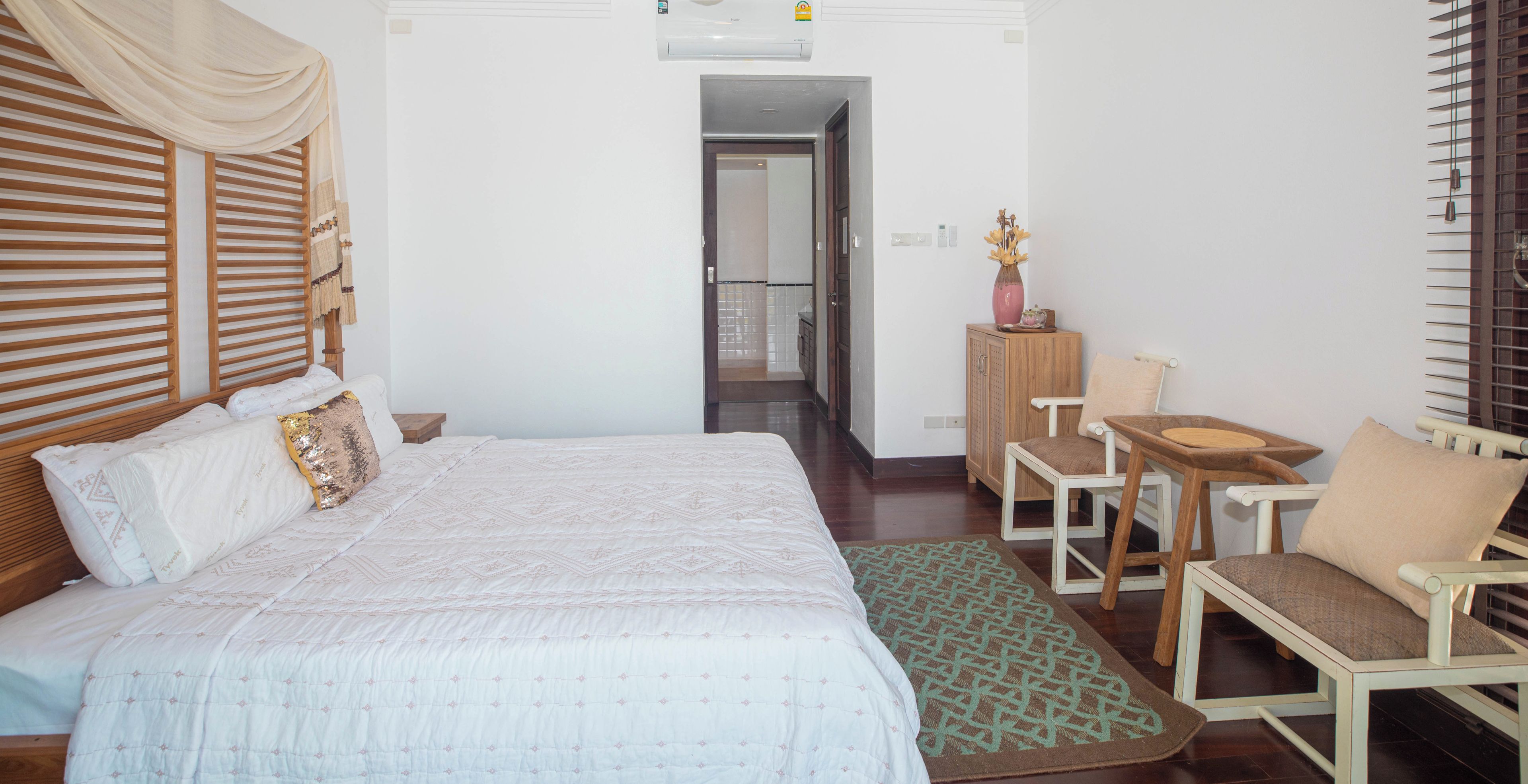 Habitación de hotel con una cama grande, cabecera de madera, una mesita con sillas, un armario y una puerta que da a otra habitación.