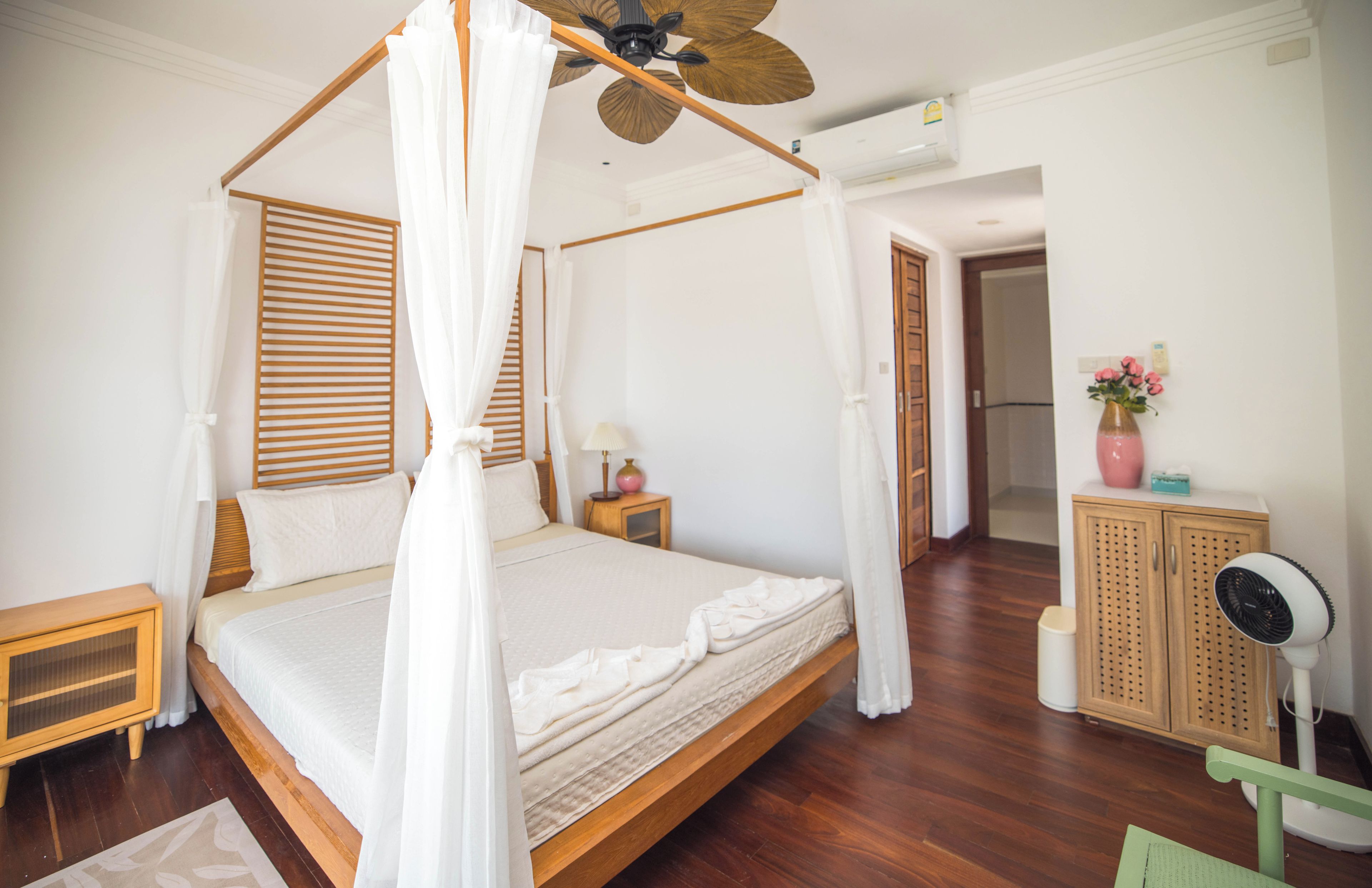 A four-poster canopy bed with a wooden headboard, a bedside table with a lamp, and a cabinet with a fan.