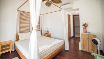 A four-poster canopy bed with a wooden headboard, a bedside table with a lamp, and a cabinet with a fan.