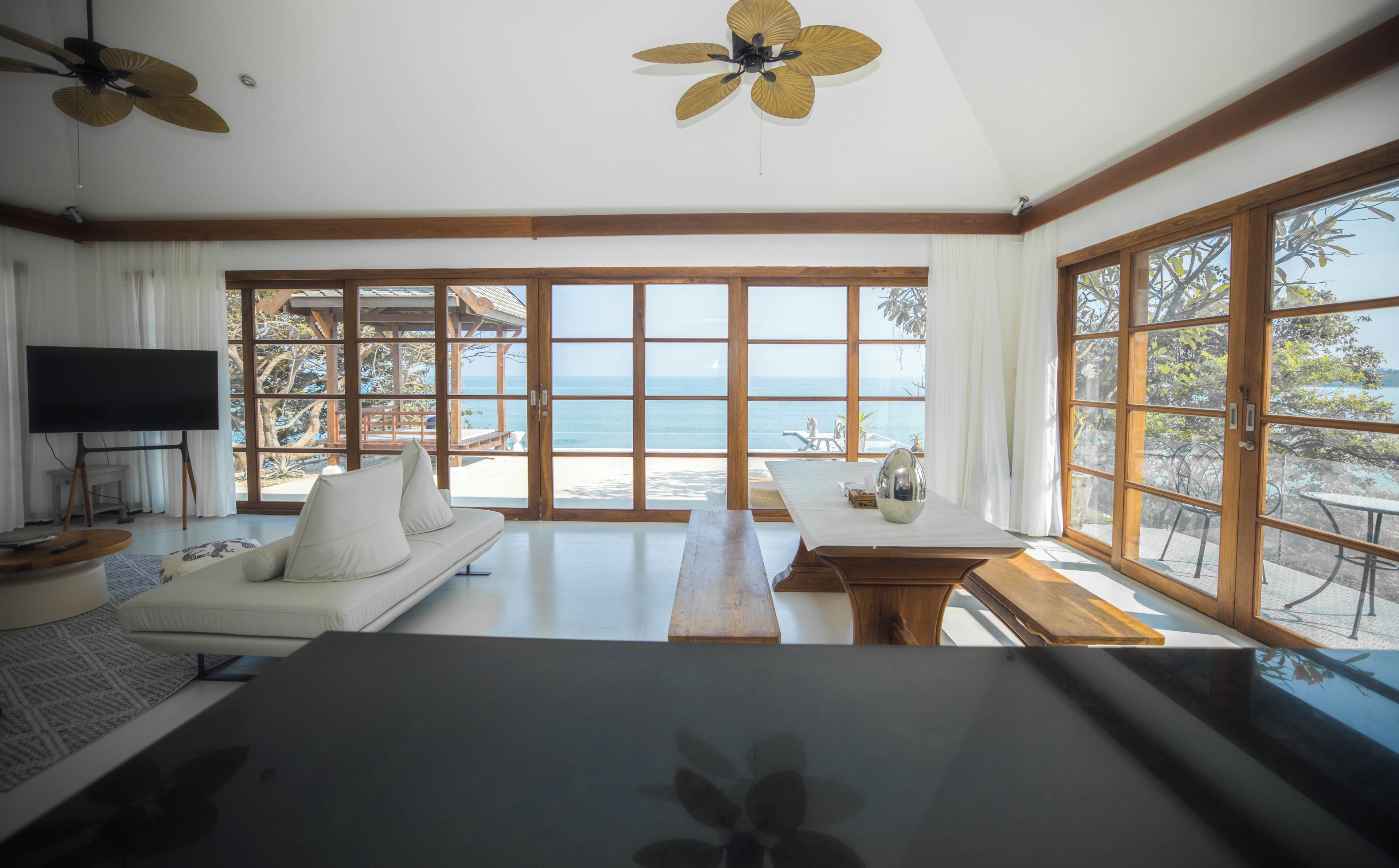 A spacious room with a large window offering a view of the sea, a sofa, a dining table, and ceiling fans.