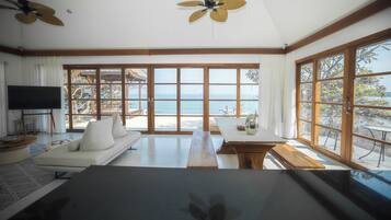 A spacious room with a large window offering a view of the sea, a sofa, a dining table, and ceiling fans.