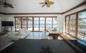 Amplia habitación con un gran ventanal que ofrece vistas al mar, un sofá, una mesa de comedor y ventiladores de techo.