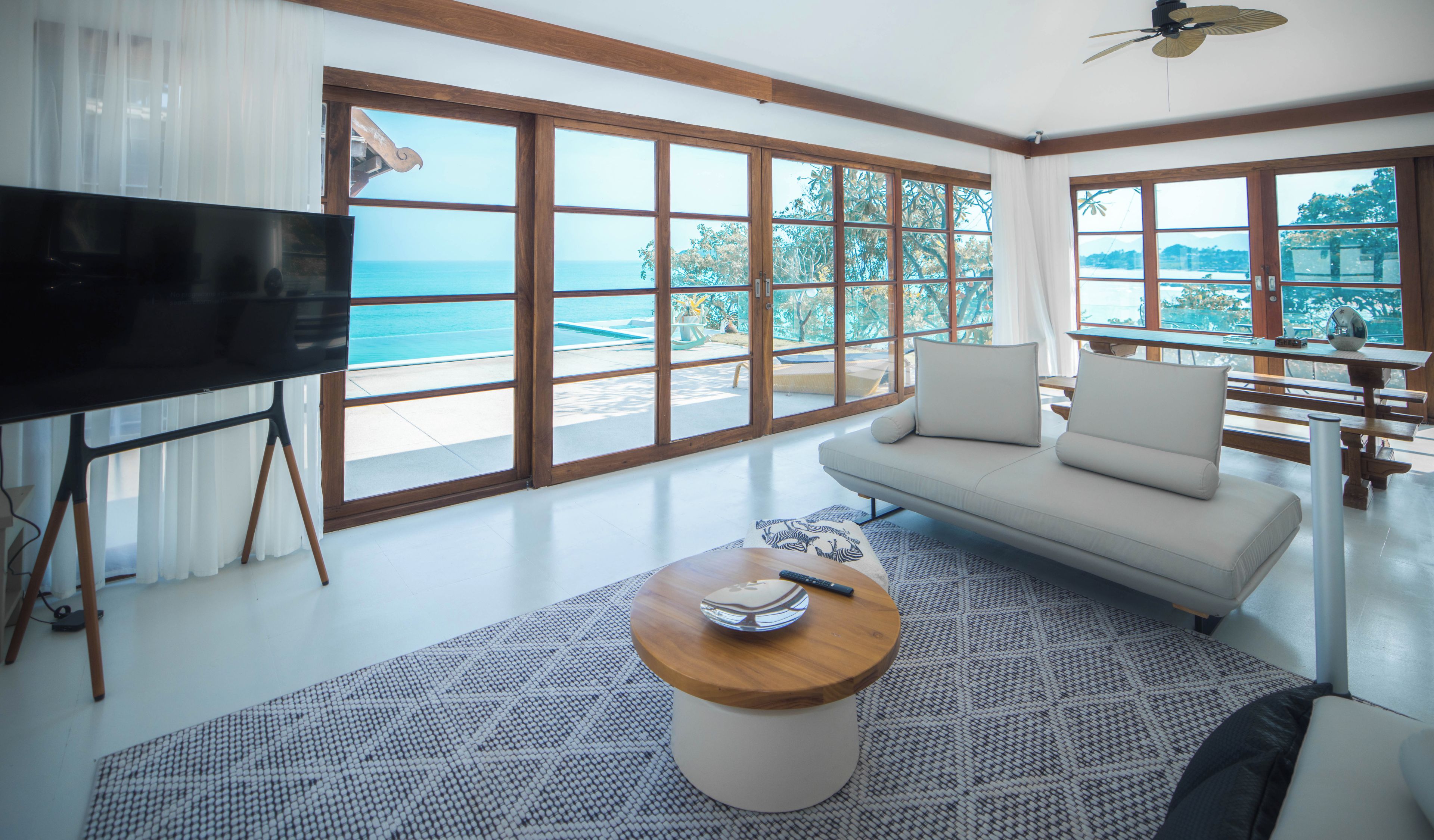 A modern living room with a large TV, a sofa, a coffee table, and a view of the ocean through expansive windows.
