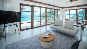 A modern living room with a large TV, a sofa, a coffee table, and a view of the ocean through expansive windows.