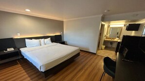 Quarto de hotel com uma cama grande, uma escrivaninha com cadeira, mesa de cabeceira e banheiro visível através de uma porta aberta.