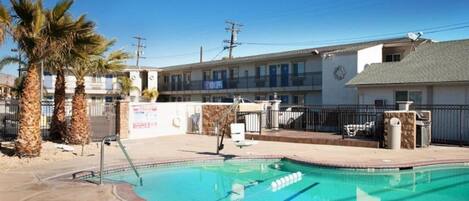 Una piscina con borde de ladrillo, palmeras y un edificio con un letrero al fondo.