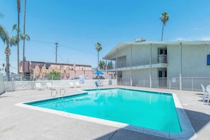 Una piscina con agua cristalina azul, rodeada por un solárium de hormigón, y un edificio con balcones al fondo.