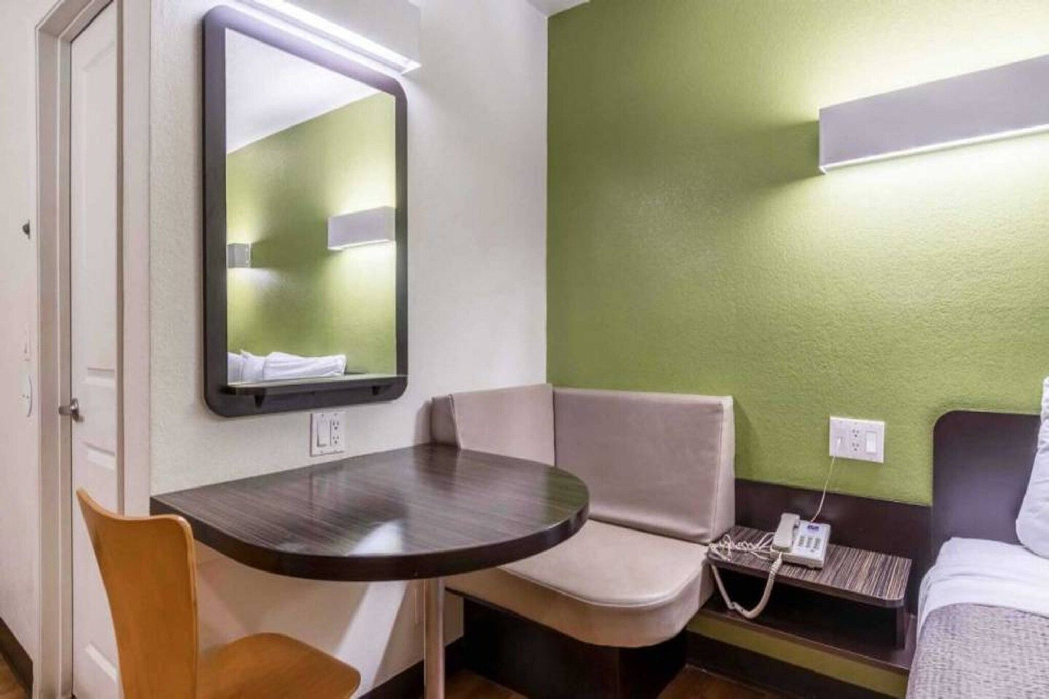 A compact hotel room with a single bed, a small table, a chair, a bedside table with a phone, and a mirror.