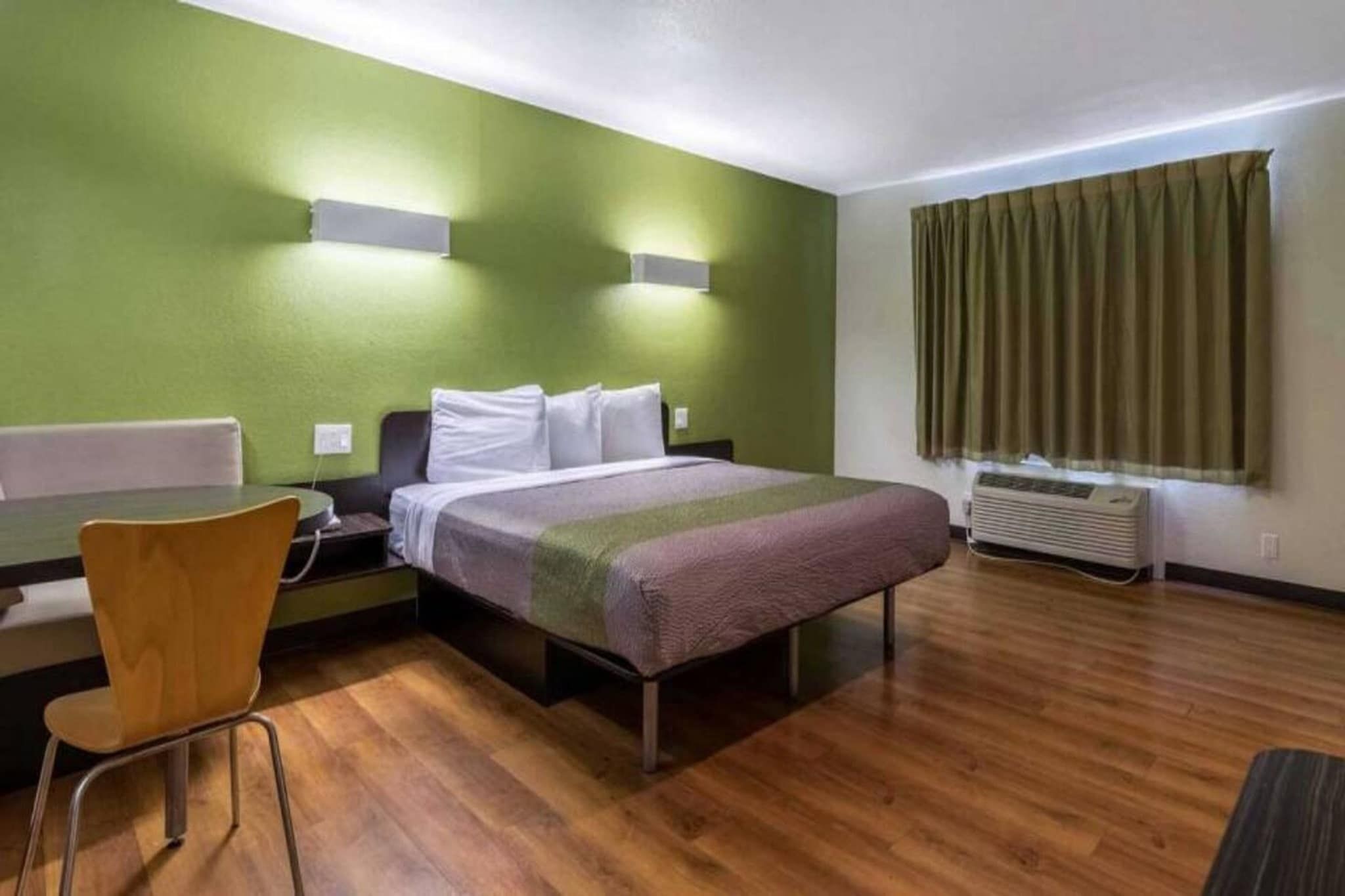 A hotel room with a bed, a desk, a chair, a window with curtains, and an air conditioning unit.