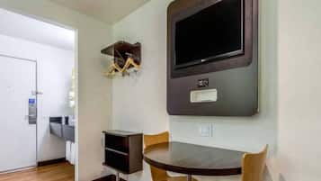 A hotel room with a dining table, chairs, a TV mounted on the wall, and a small shelf with hanging items.