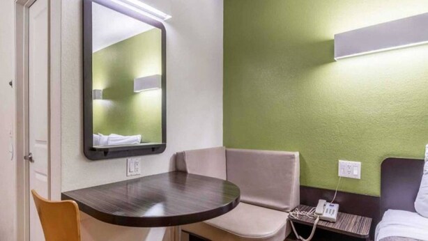 Una camera d'albergo modesta con un tavolino rotondo, una sedia, un letto, un comodino con un telefono e uno specchio fissato alla parete.
