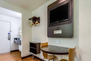 Une chambre d’hôtel équipée d’une table à manger, de chaises, d’un téléviseur fixé au mur et d’une petite étagère où sont suspendus divers objets.