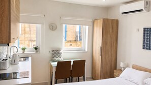 A compact bedroom with a bed, a desk with two chairs, a wardrobe, and a window with a view of a building. - Marina Studions (Bormla)