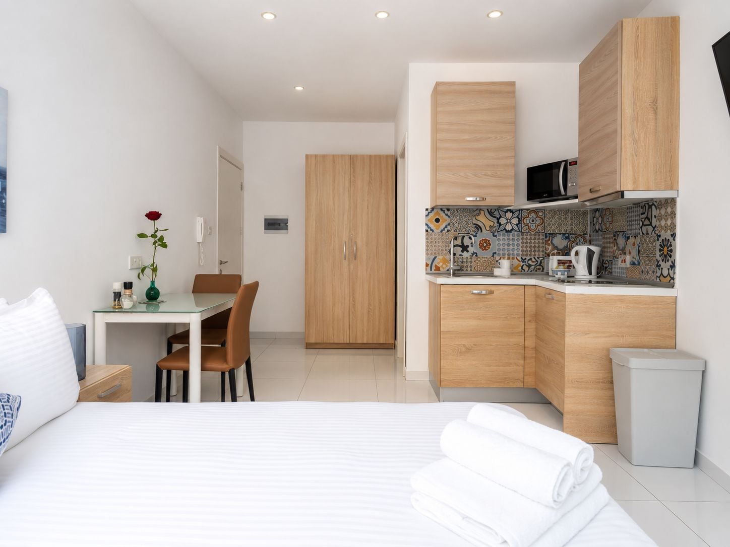 A modern hotel room with a bed, a dining table with chairs, a kitchenette with a microwave, and a cabinet.