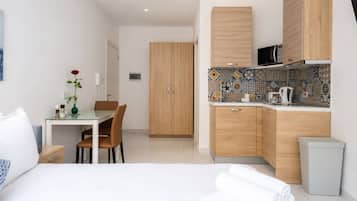 A modern hotel room with a bed, a dining table with chairs, a kitchenette with a microwave, and a cabinet.