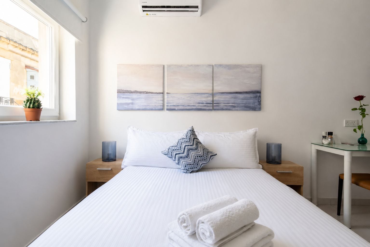 A hotel room with a bed, a nightstand, a desk, a potted plant, and a painting on the wall.