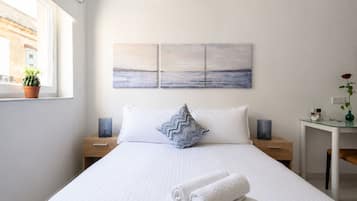 A hotel room with a bed, a nightstand, a desk, a potted plant, and a painting on the wall.