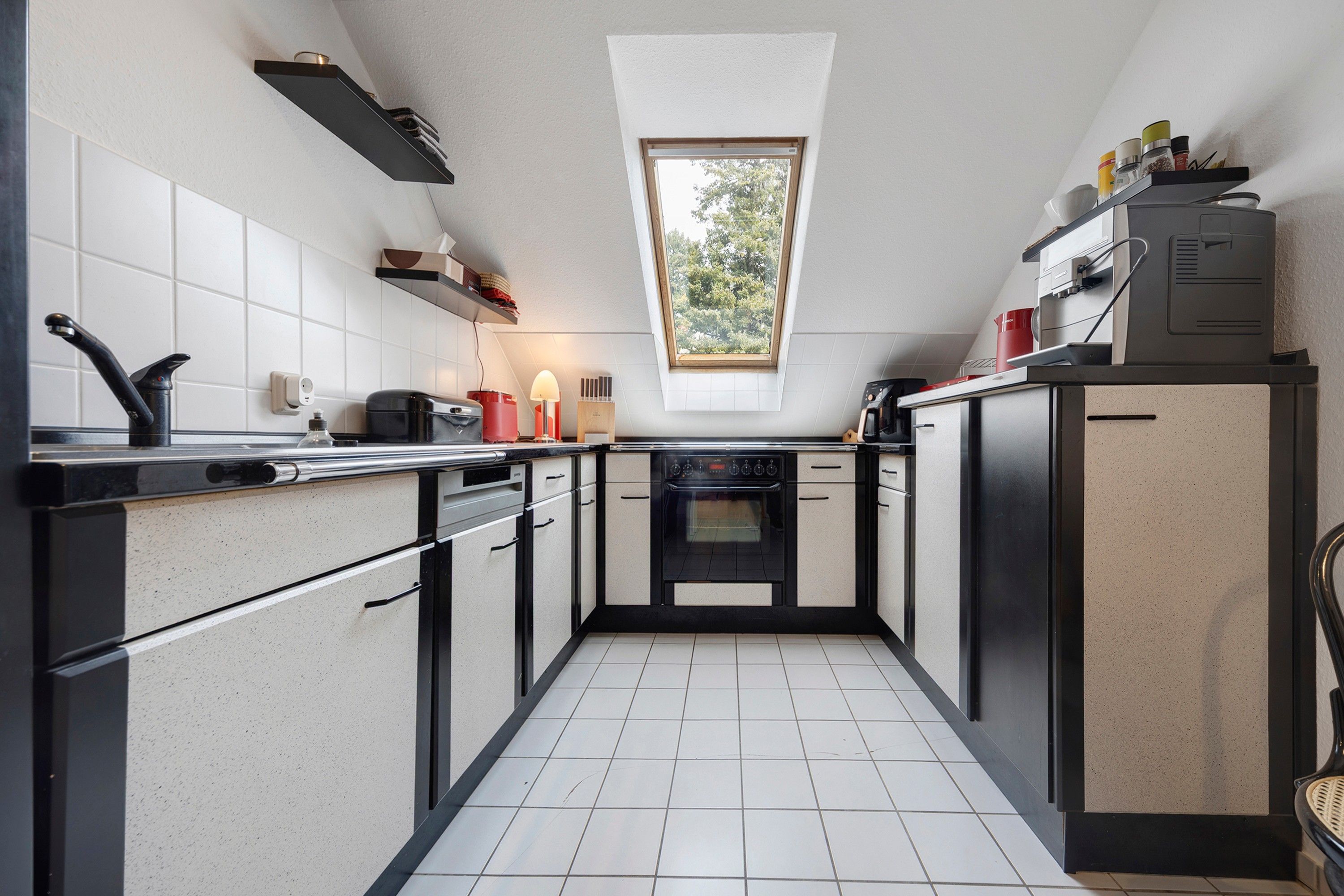 Et kompakt køkken med ovenlys, hvide flisegulv og moderne hårde hvidevarer.