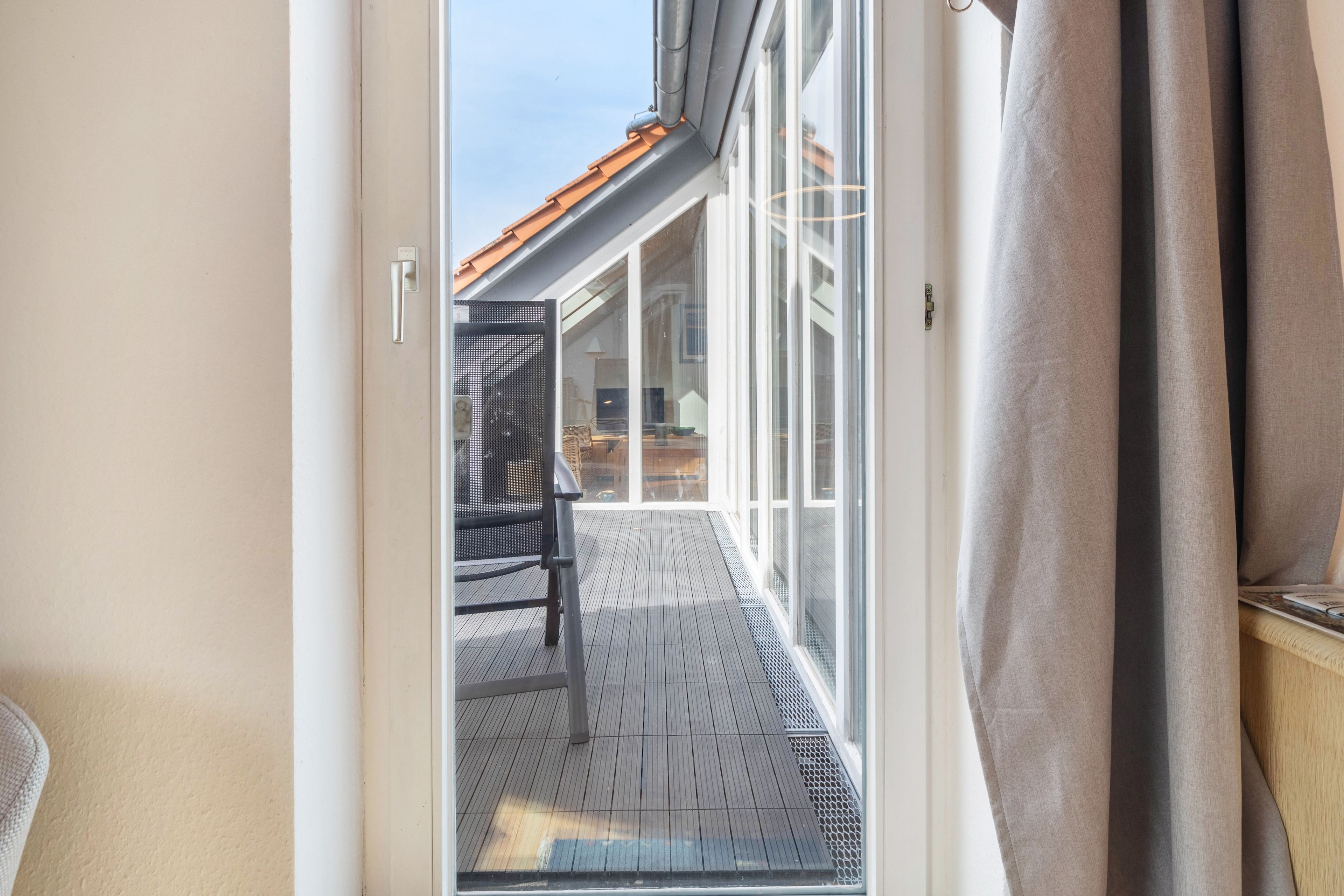 Uitzicht door een glazen deur op een houten terras met een stoel en een tafel, dat uitkomt bij een gebouw met een pannendak.