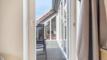 Uitzicht door een glazen deur op een houten terras met een stoel en een tafel, dat uitkomt bij een gebouw met een pannendak.