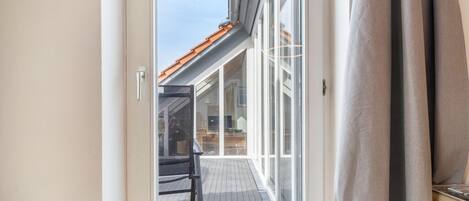 A view through a glass door onto a wooden deck with a chair and a table, leading to a building with a tiled roof.