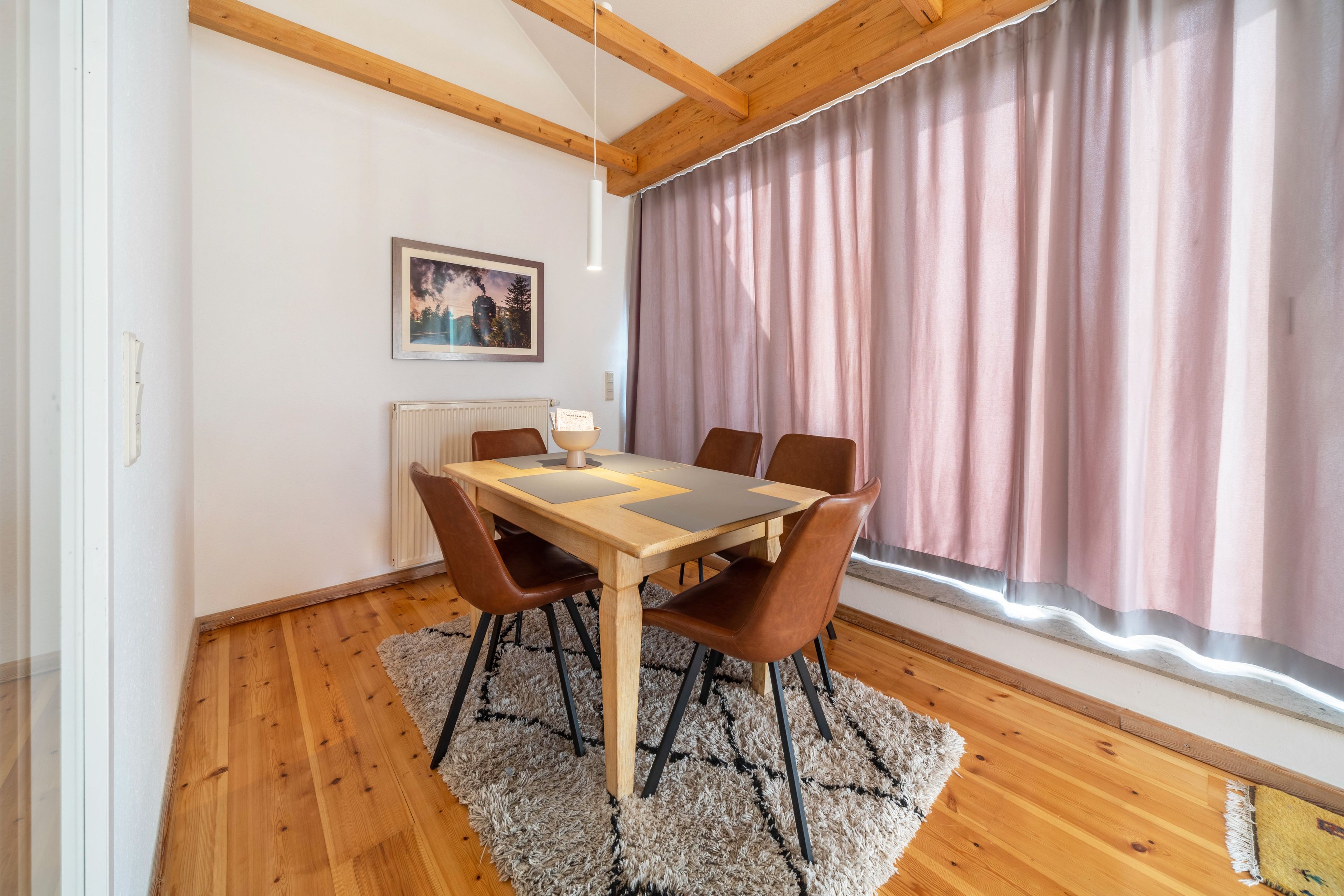 Ein Essbereich mit Holztisch, sechs Stühlen, einem Teppich, einem Bild an der Wand und einem Fenster mit Vorhängen.