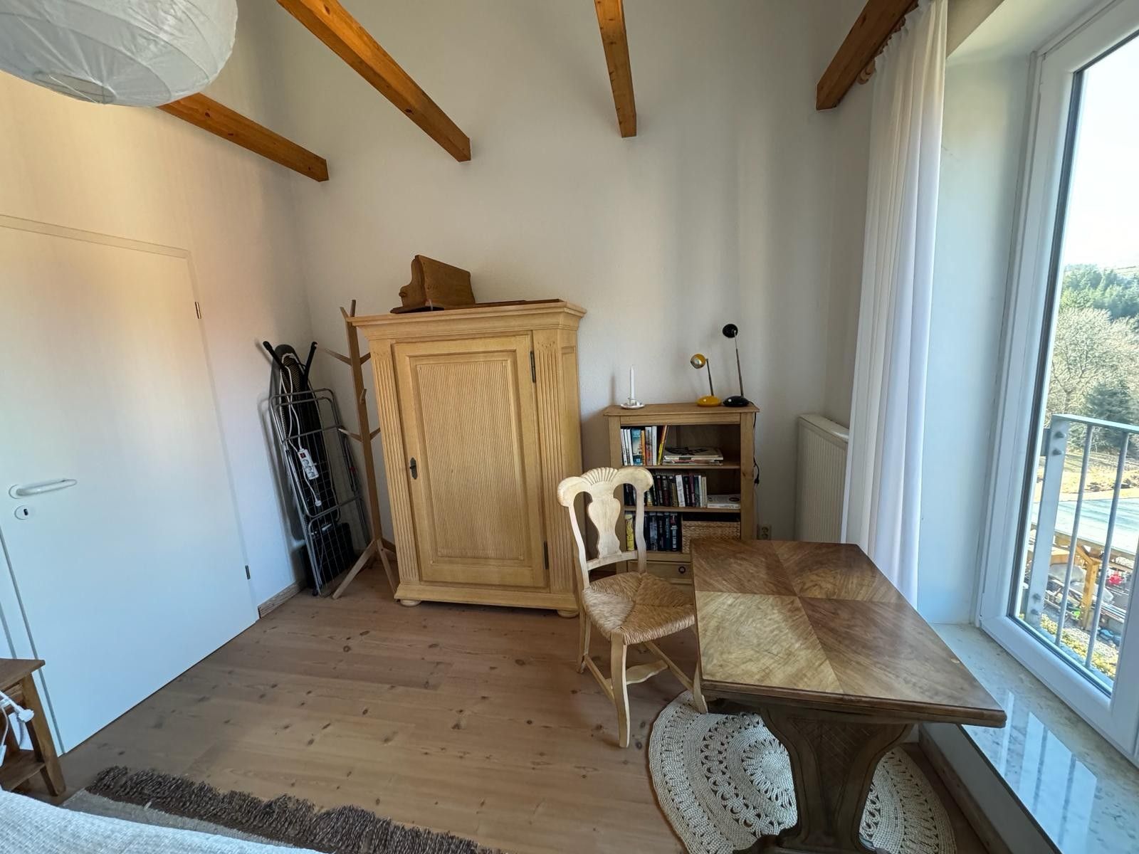 Ein Zimmer mit Holzboden, einem Holztisch mit einem gewebten Stuhl, einem Schrank und einem Bücherregal.