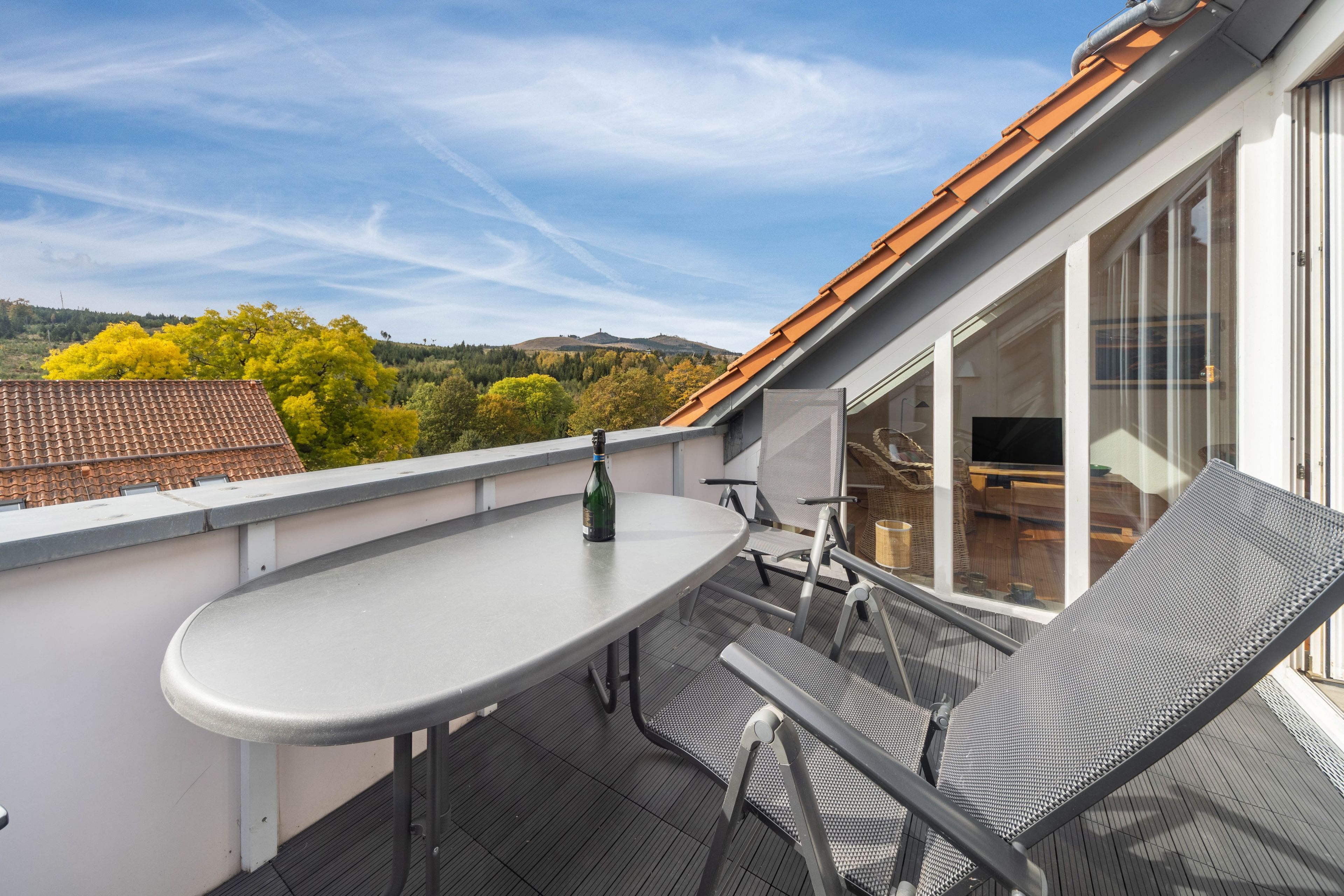 Eine Dachterrasse mit Tisch und Stühlen, einer Flasche und Blick auf Bäume und Hügel.