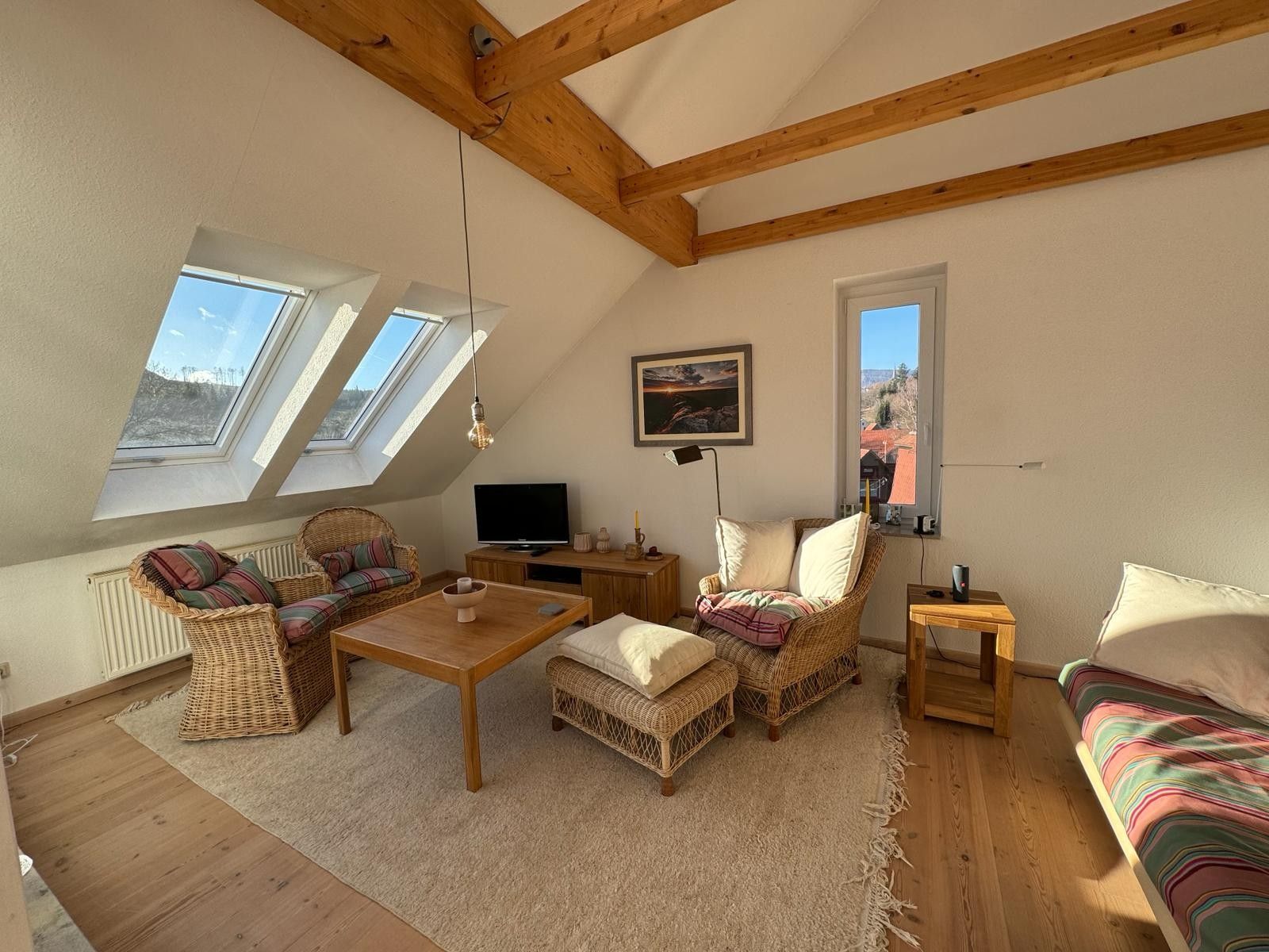 Ein gemütliches Schlafzimmer im Loft-Stil mit schrägem Dach, Holzbalken, einem großen Fenster, einem Bett, einem Fernseher und einer Essecke mit Korbmöbeln.