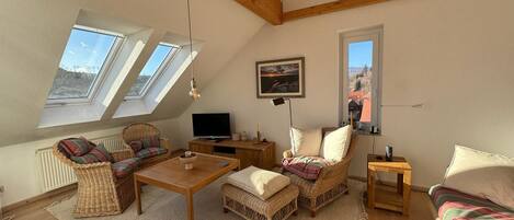 A cozy loft bedroom with a sloped ceiling, wooden beams, a large window, a bed, a TV, and a dining area with wicker furniture.