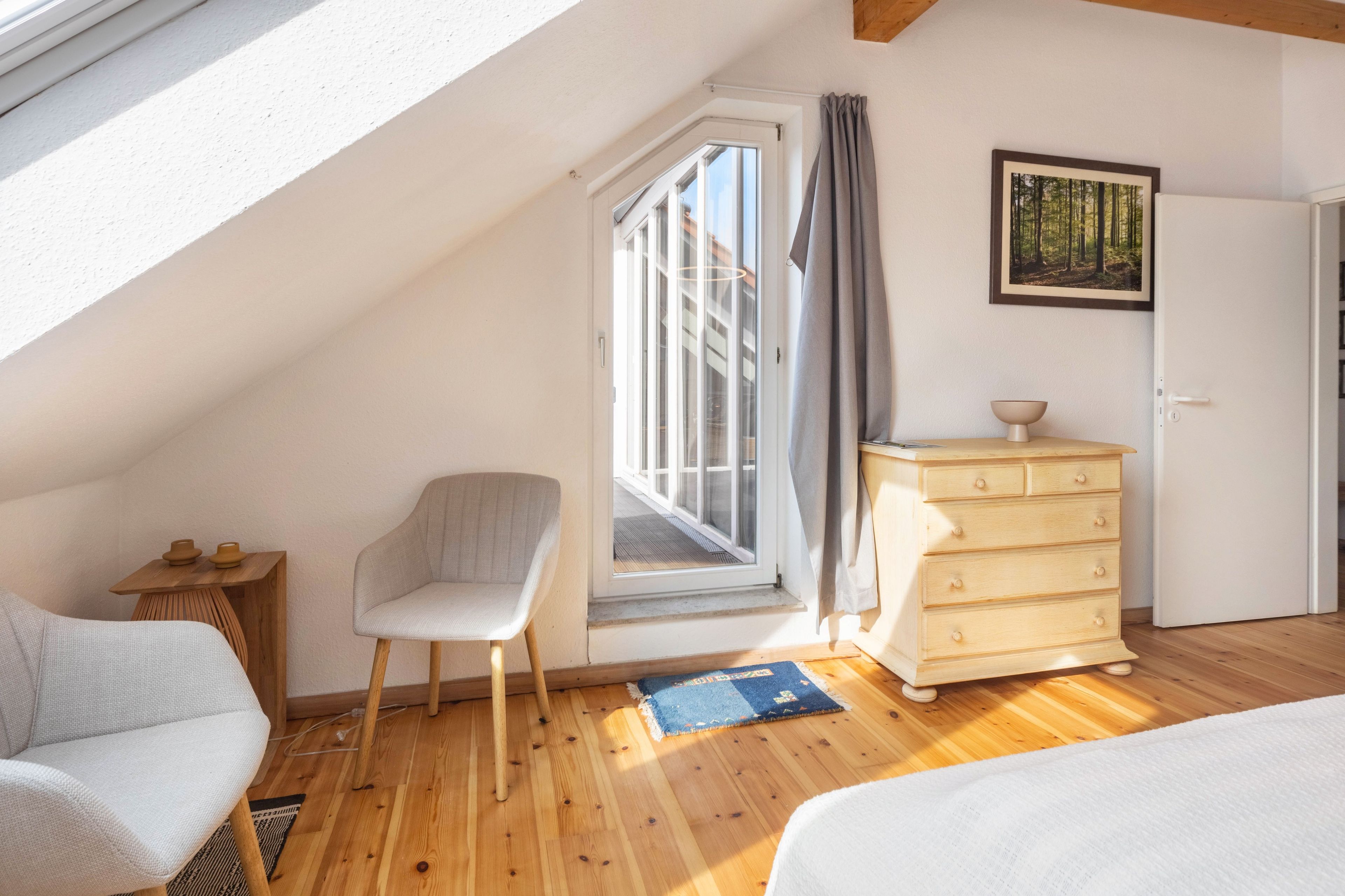 Ein Schlafzimmer mit schrägen Wänden, einem Fenster mit Vorhängen, einem Sessel, einer Kommode und einem Bett.