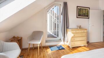 Ein Schlafzimmer mit schrägen Wänden, einem Fenster mit Vorhängen, einem Sessel, einer Kommode und einem Bett.