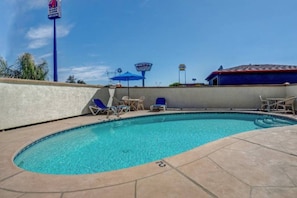 Piscina com espreguiçadeiras e guarda-sóis, cercada por um muro baixo e um edifício com telhado vermelho.