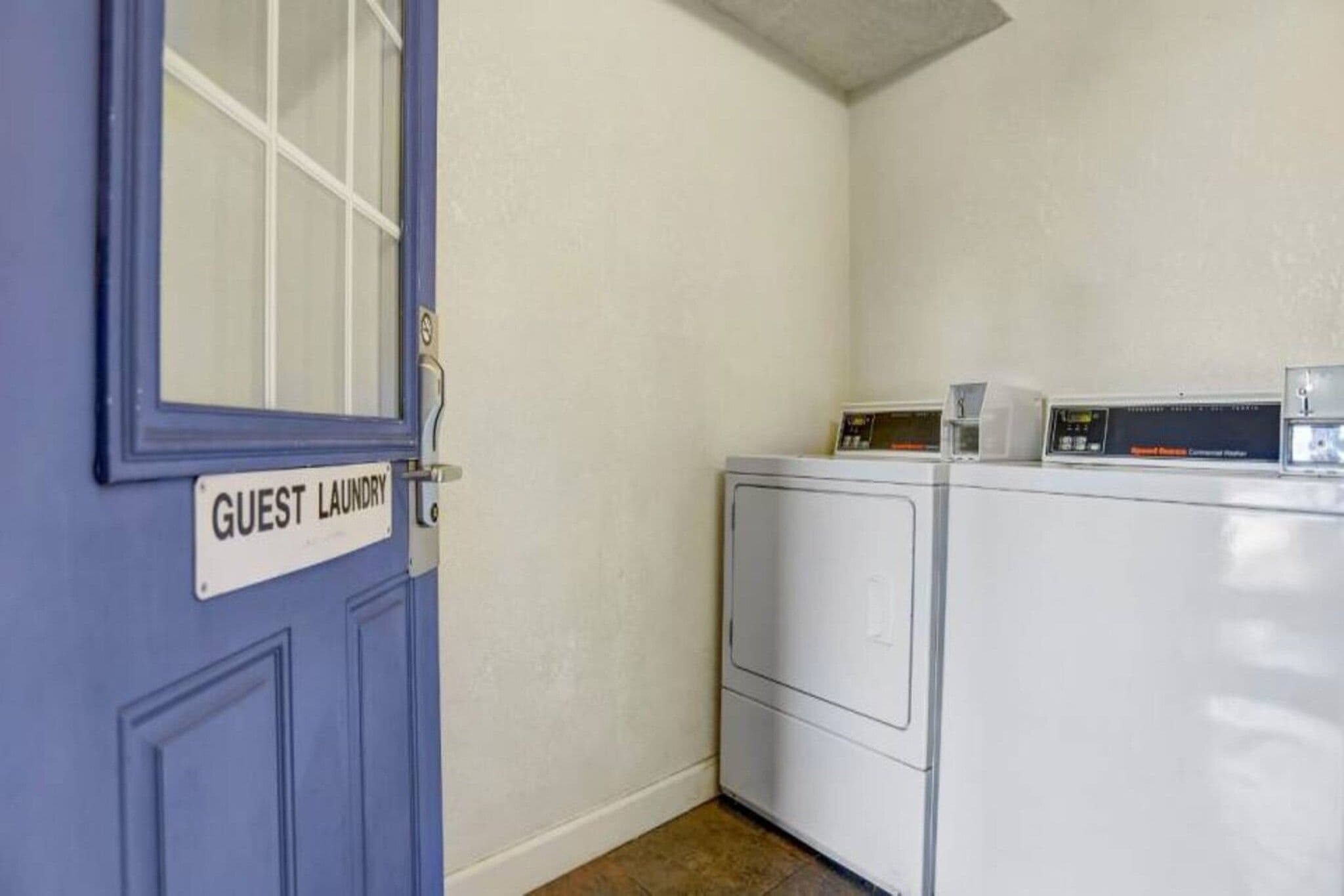 Un cuarto de lavandería con una puerta azul que indica 'LAVANDERÍA DE HUÉSPEDES' y una lavadora y secadora de carga frontal.