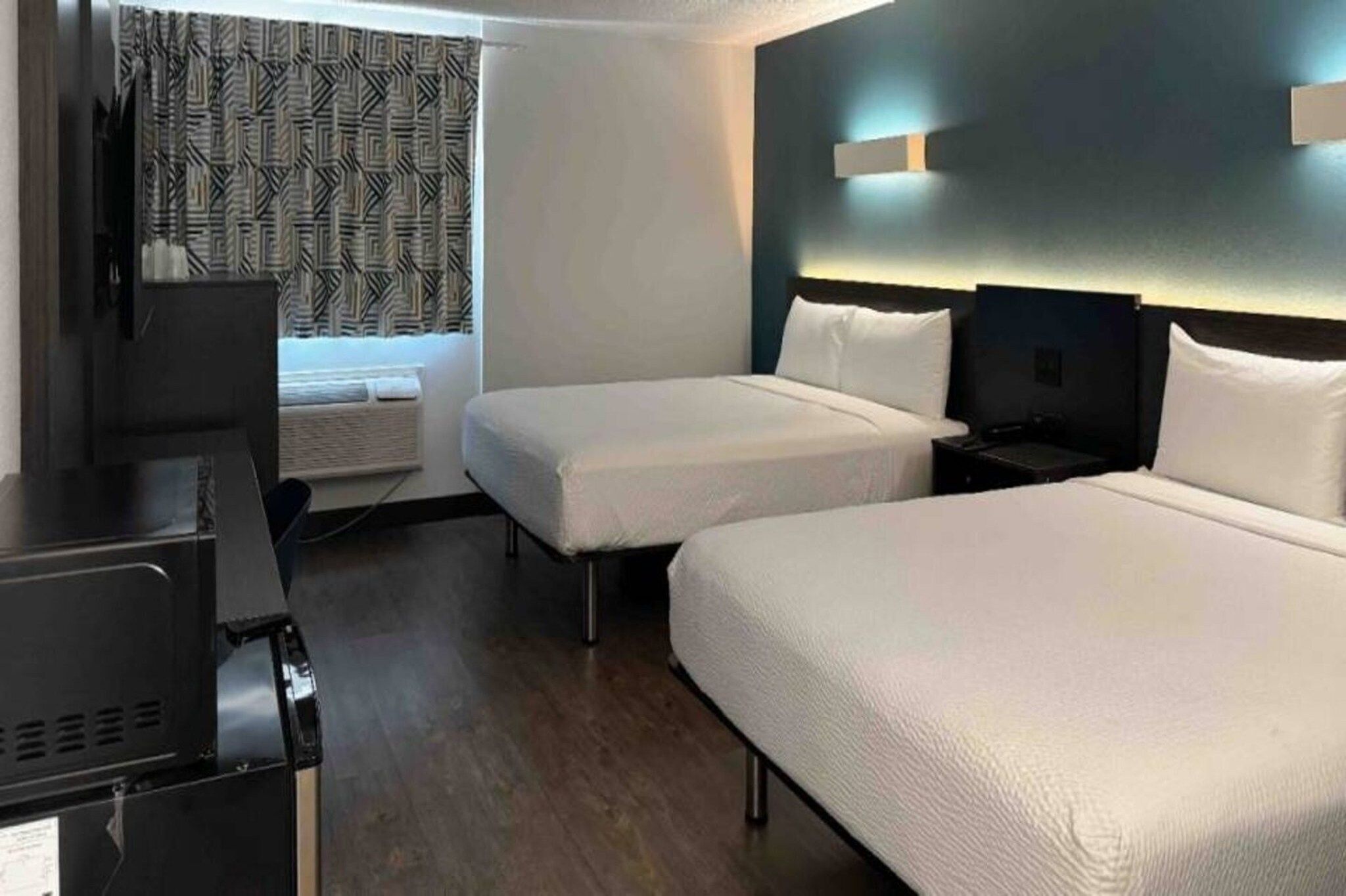 Habitació d'hotel amb dos llits individuals, una taula de treball amb un ordinador, un televisor i una finestra amb cortines estampades.