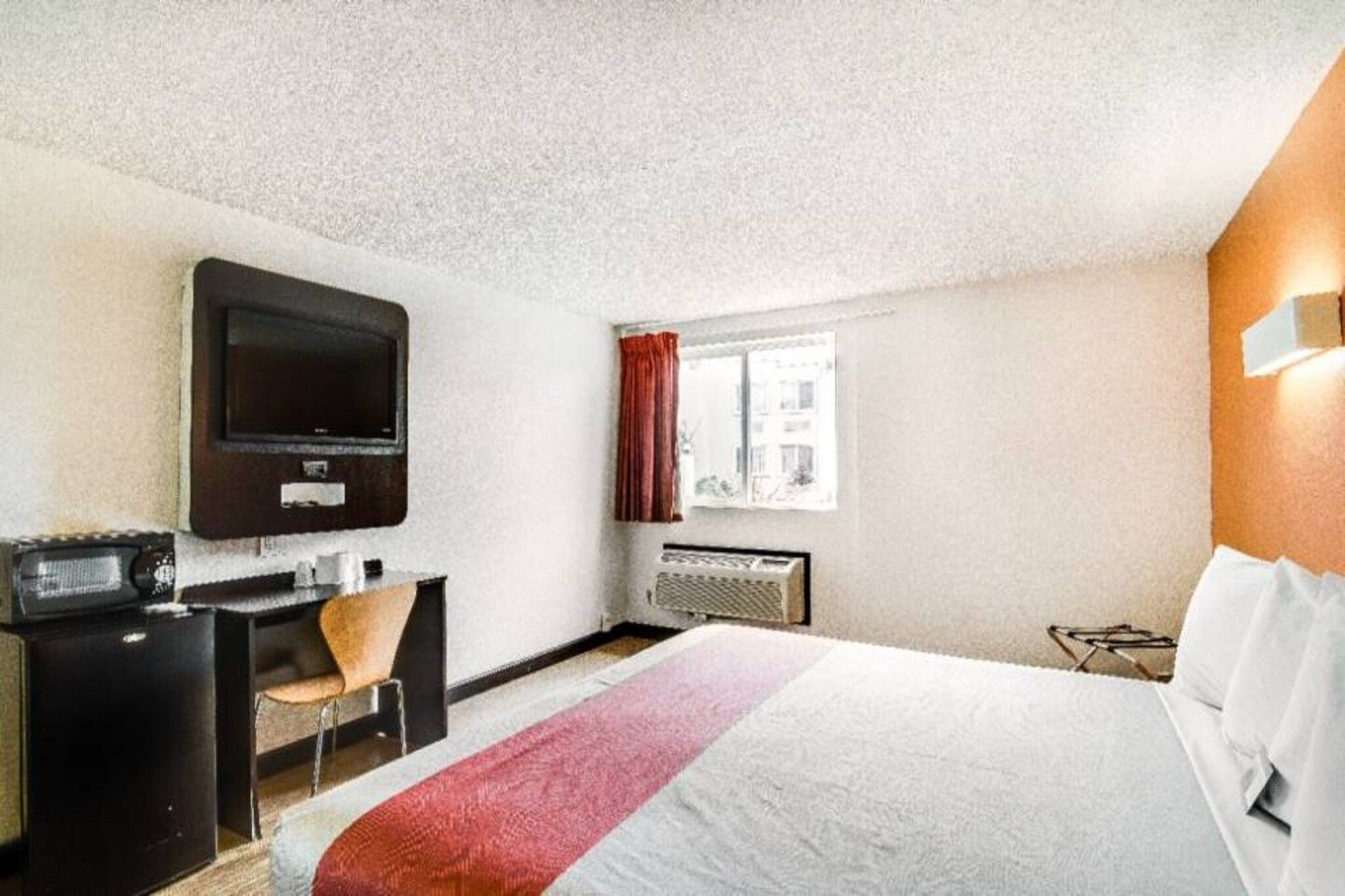 A hotel room with a bed, a desk, a television, a window with curtains, and a radiator.