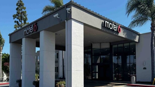 Um motel com entrada coberta, estacionamento e uma palmeira ao fundo.