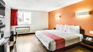 Quarto de hotel com cama, escrivaninha, cadeira, janela com cortinas e luminária na parede.