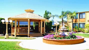 A gazebo with a tiled roof, surrounded by a circular flower bed with various flowers, and a building with multiple windows in the background.