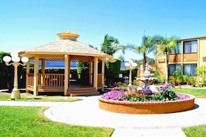 Un kiosque surmonté d’un toit en tuiles, entouré d’un parterre circulaire où poussent différentes espèces de fleurs, et un bâtiment avec de nombreuses fenêtres en arrière-plan.