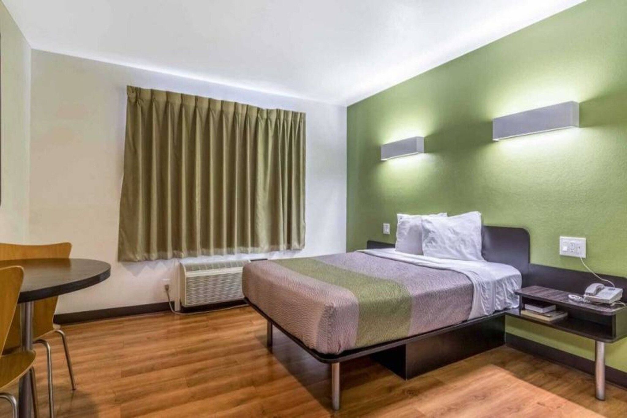 A hotel room with a bed, a desk, a chair, a window with curtains, and a wall-mounted air conditioner.