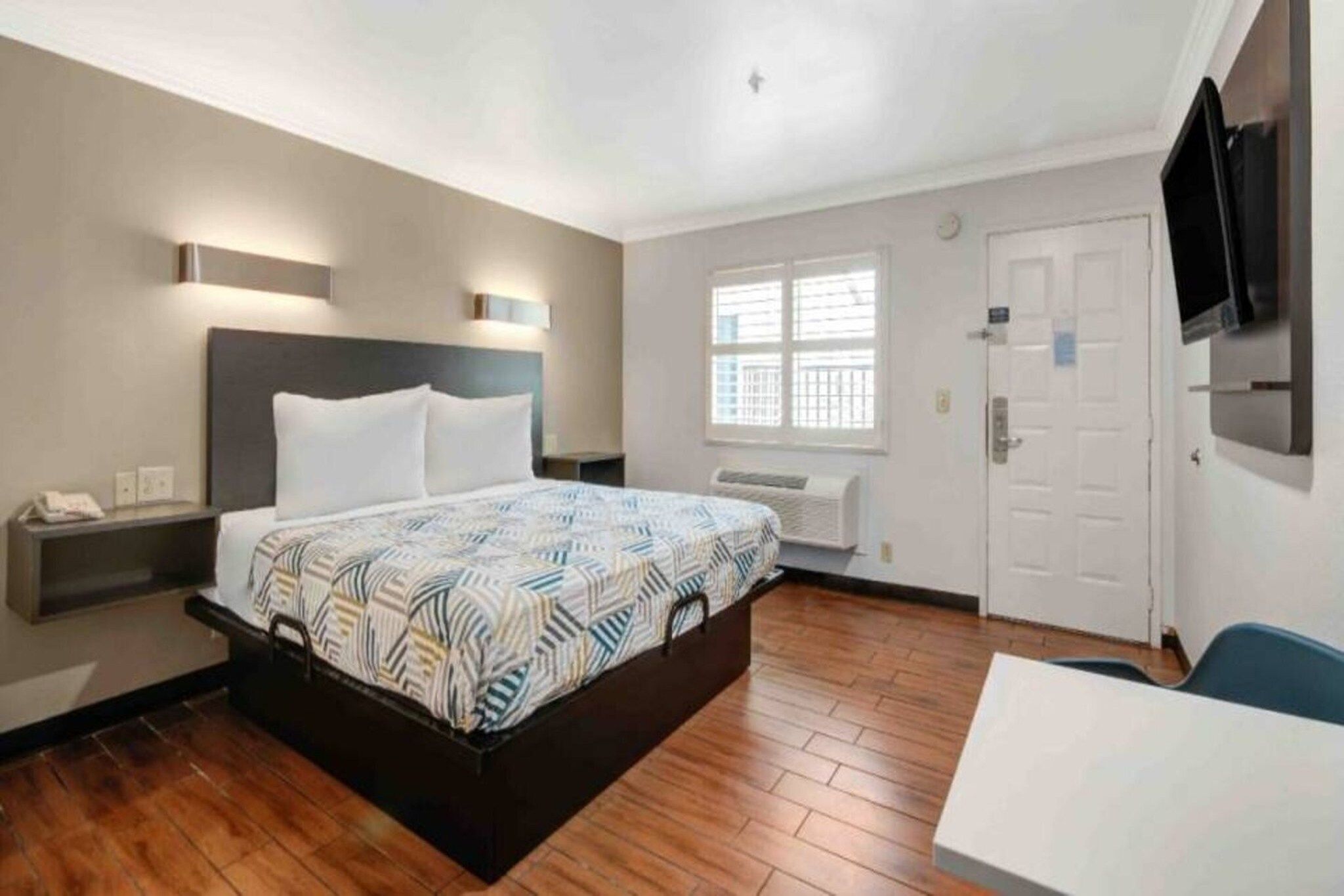 A hotel room with a bed, bedside table, wall-mounted TV, window, and a door.