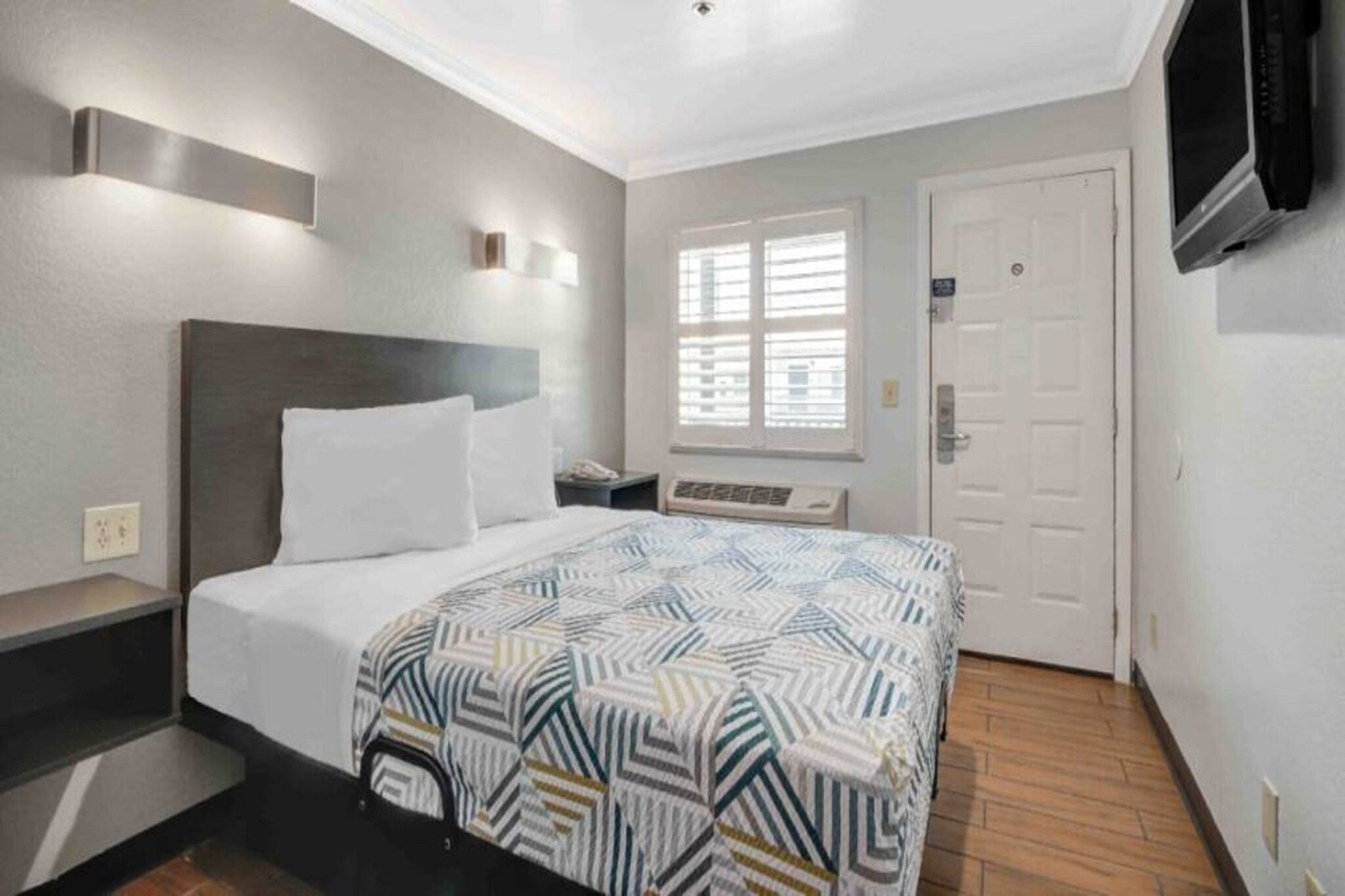 A hotel room with a bed, bedside table, a window with blinds, a door, and a television mounted on the wall.