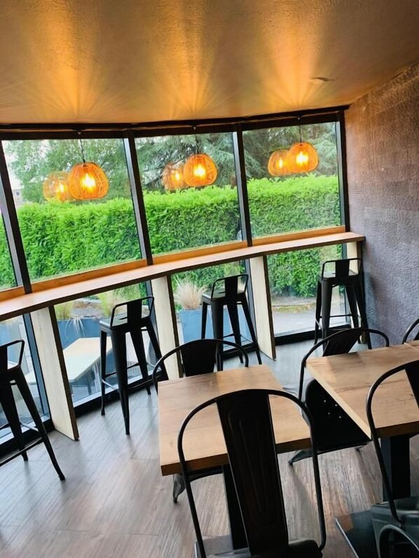 An indoor dining area with tables and chairs, large windows, and hanging pendant lights. - LE MOKCA (Meylan)