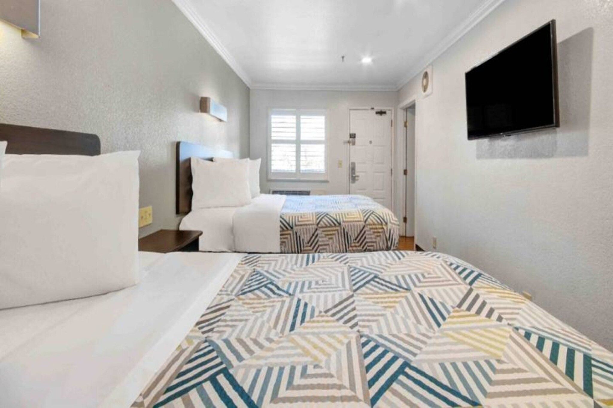 A hotel room with two beds, a television mounted on the wall, and a window providing natural light.