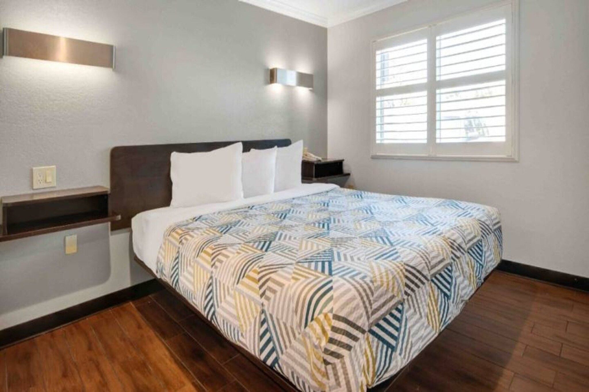 A hotel room with a bed, a nightstand, a wall-mounted light, and a window with blinds.
