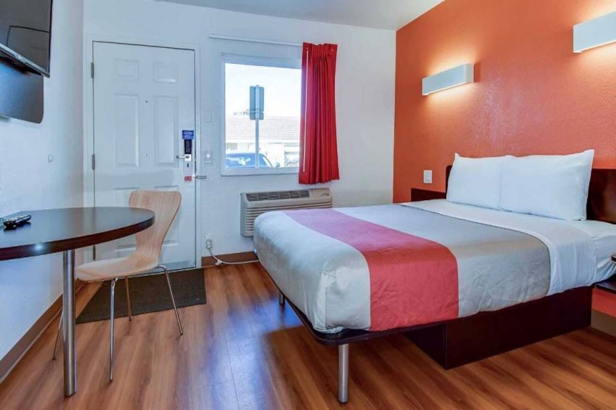 A hotel room with a bed, a desk, a chair, a window with curtains, and a wall-mounted air conditioning unit.